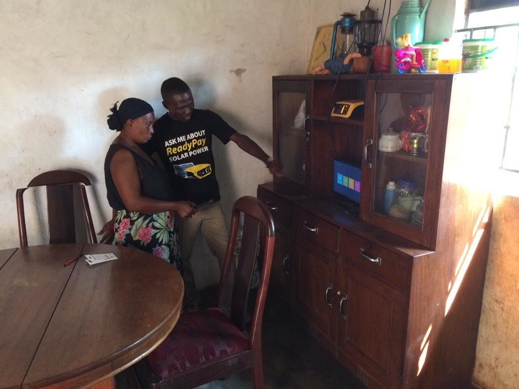 A sales agent for Fenix International visits a customer in her farmhouse in rural Uganda. The yellow box on the bookshelf is the battery for her solar system. 